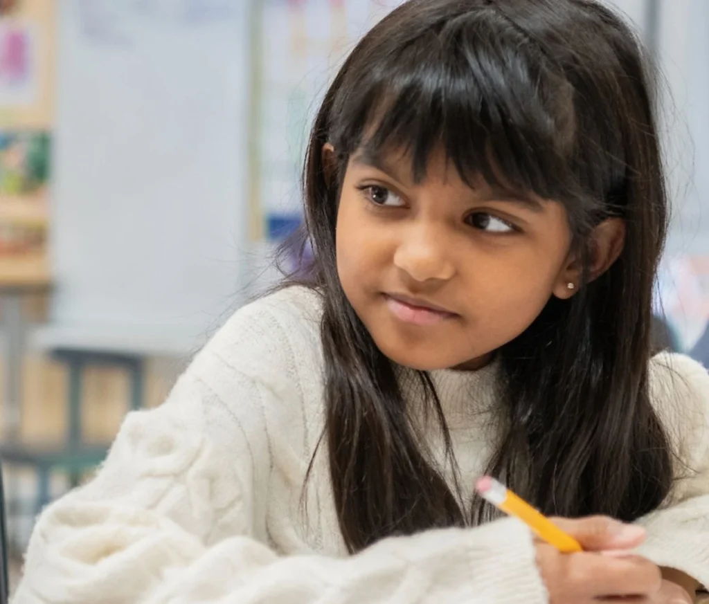Child in classroom holding pencil, wearing sweater.