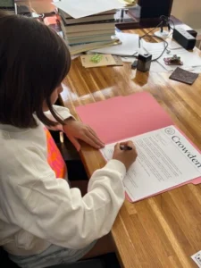 Child signing document at wooden desk.