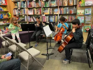 Children play string instruments in a library setting.