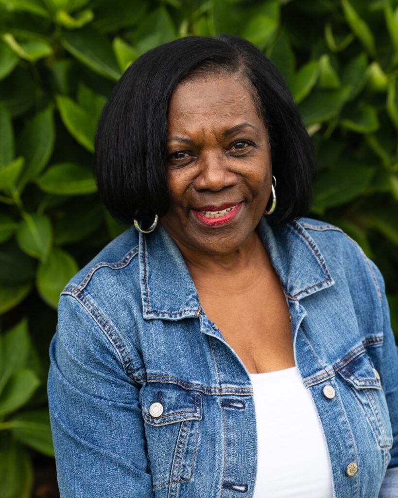 Smiling woman in denim jacket with greenery background.
