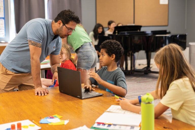 Teacher helps children with laptop in classroom.