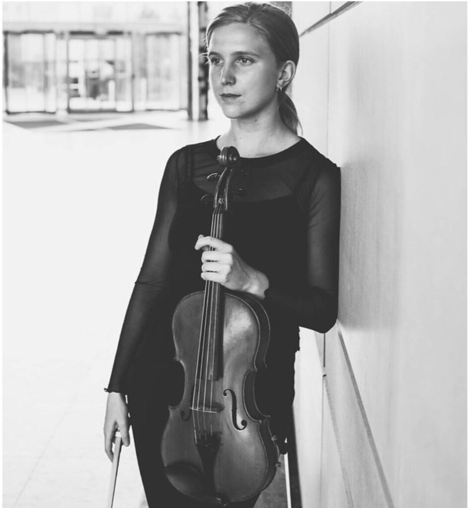 Person holding a violin, leaning against a wall.