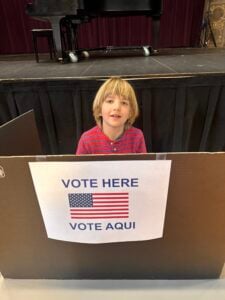 Child in a voting booth with sign 'Vote Here'