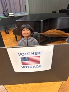 Child seated at voting booth with sign.