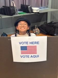 Child smiling at voting station with sign.