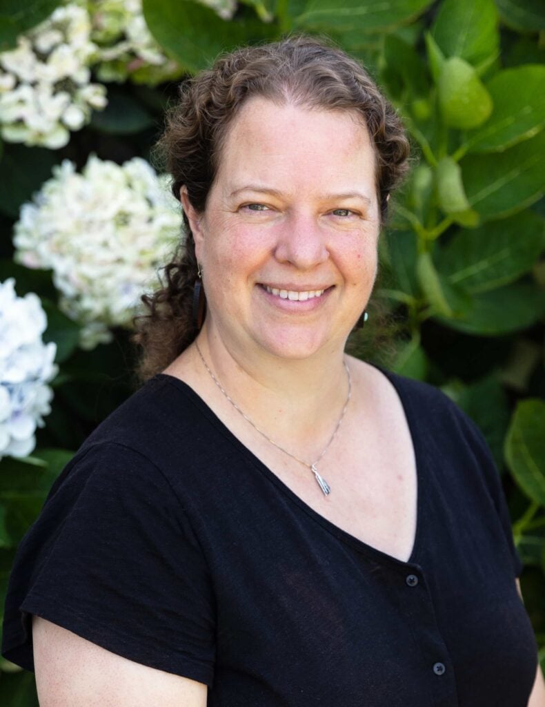 Smiling woman in black shirt, green foliage background.