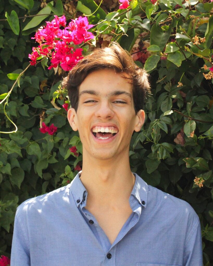 Person smiling in front of vibrant flowers and leaves.
