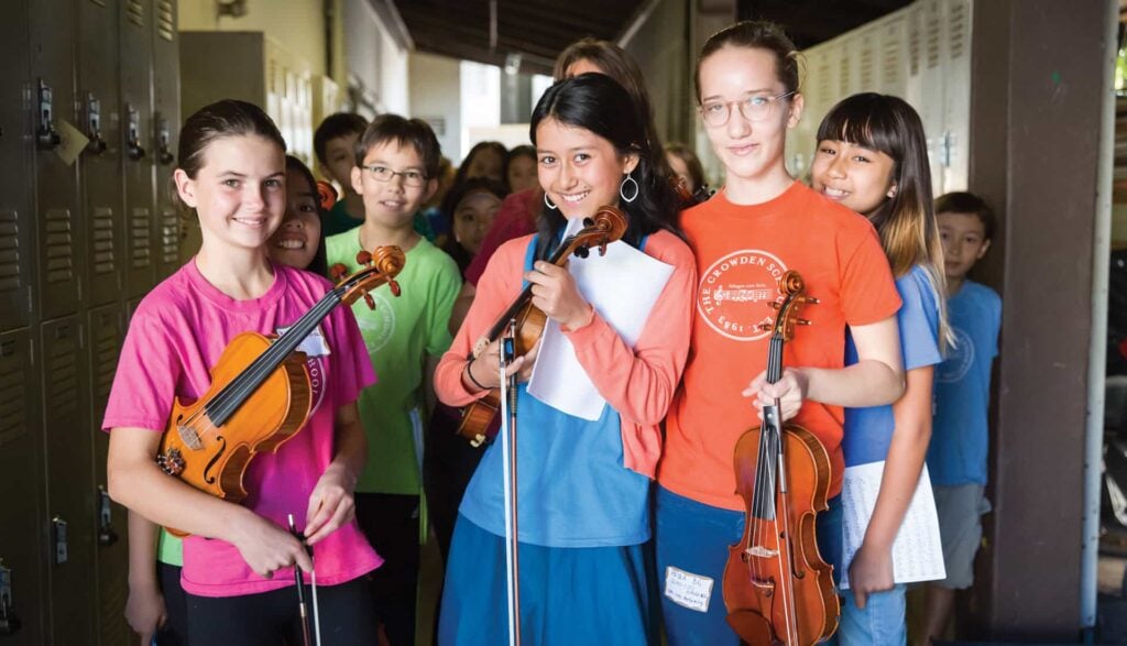 Group of children holding violins in hallway.