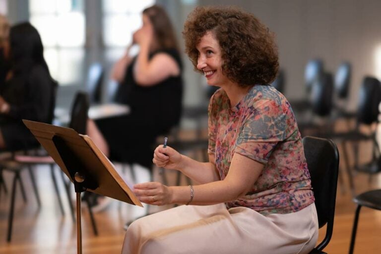 Woman seated, smiling, holding pen in rehearsal.