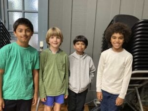 Four kids smiling in a room with chairs.