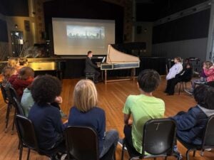 Man plays harpsichord to seated children in auditorium.