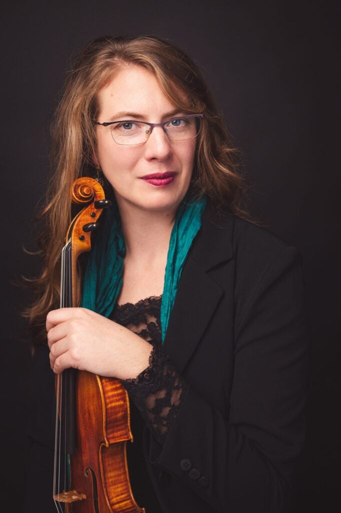 Person holding violin, wearing glasses and teal scarf.