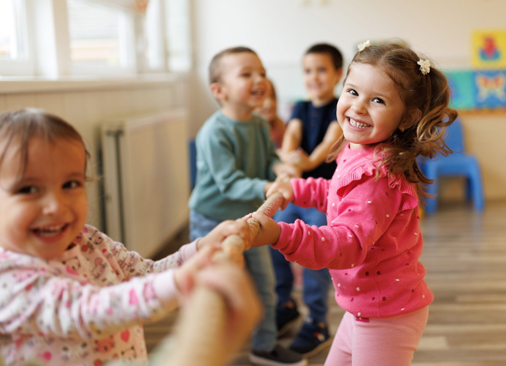 Children play tug of war indoors, smiling and happy.