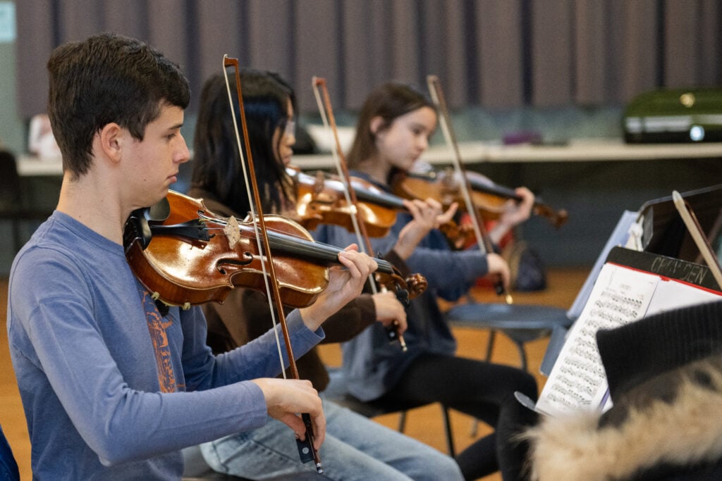 Violinists perform in a blurred, reflected orchestral scene